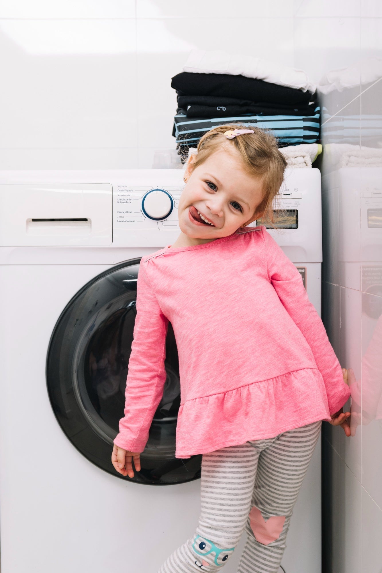 Alt text: Smiling young girl in a pink top and striped pants standing playfully in front of a washing machine with folded laundry stacked on top.