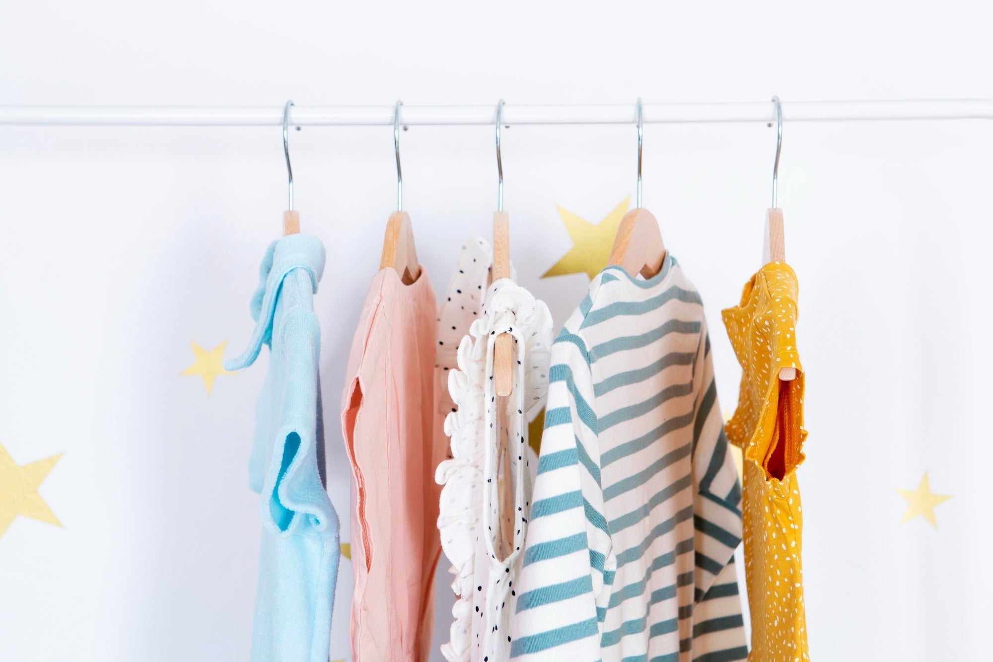 Alt text: Five colorful baby outfits hanging on wooden hangers, including ruffled, striped, and dotted designs, displayed on a white rack with star decor.