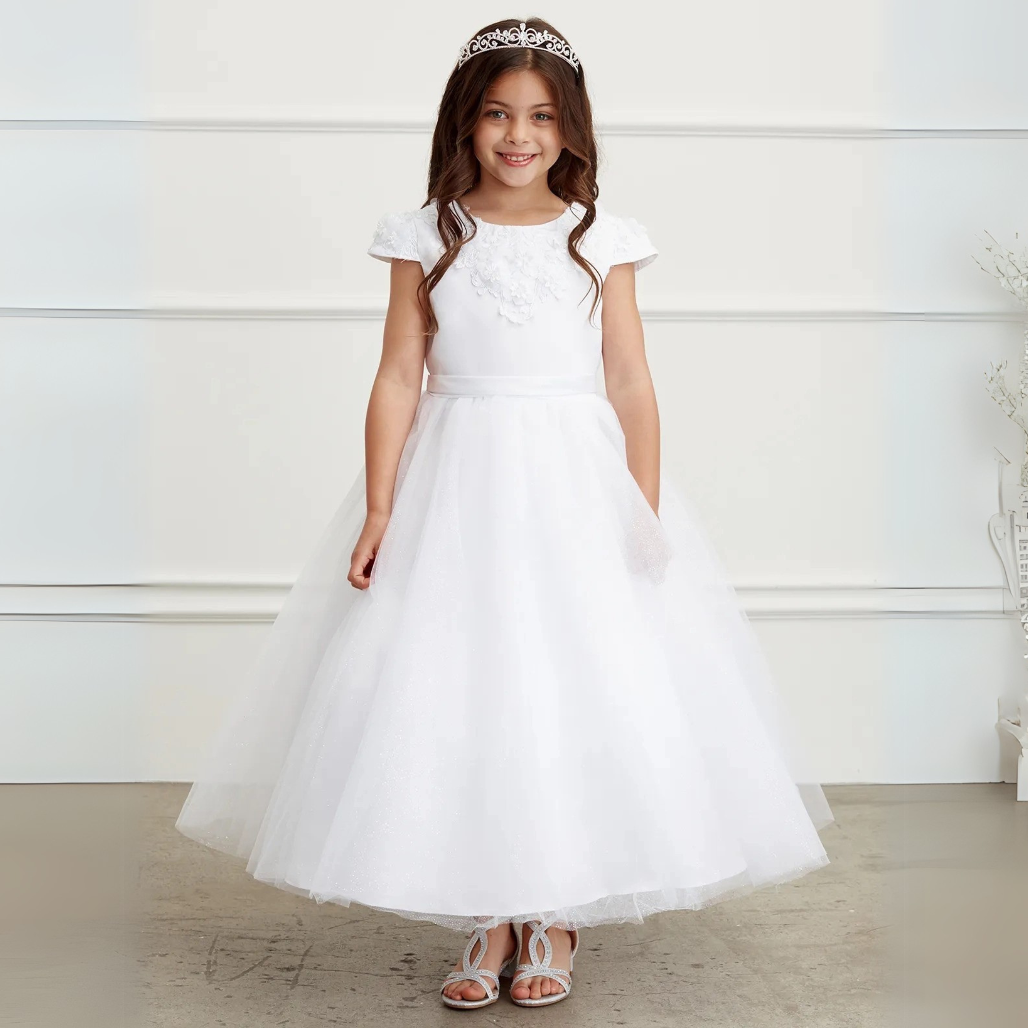 Young girl wearing a white first communion dress with tiara and sandals, standing indoors against a white wall.