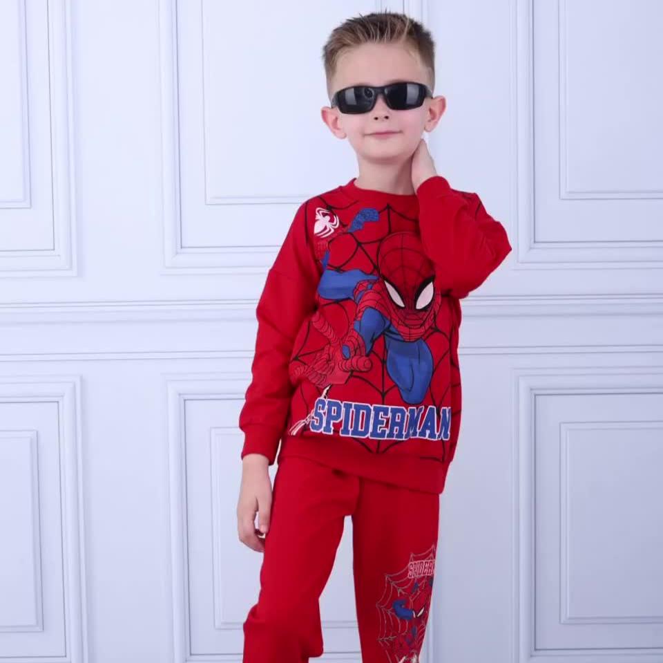 Confident young boy wearing a red Spiderman-themed sweatshirt & matching joggers, accessorized with black sunglasses, posing in front of a white wall.