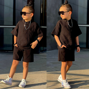 Stylish young boy wearing a black textured short-sleeve shirt & matching shorts, accessorized with oversized sunglasses, jewelry, and white sneakers.