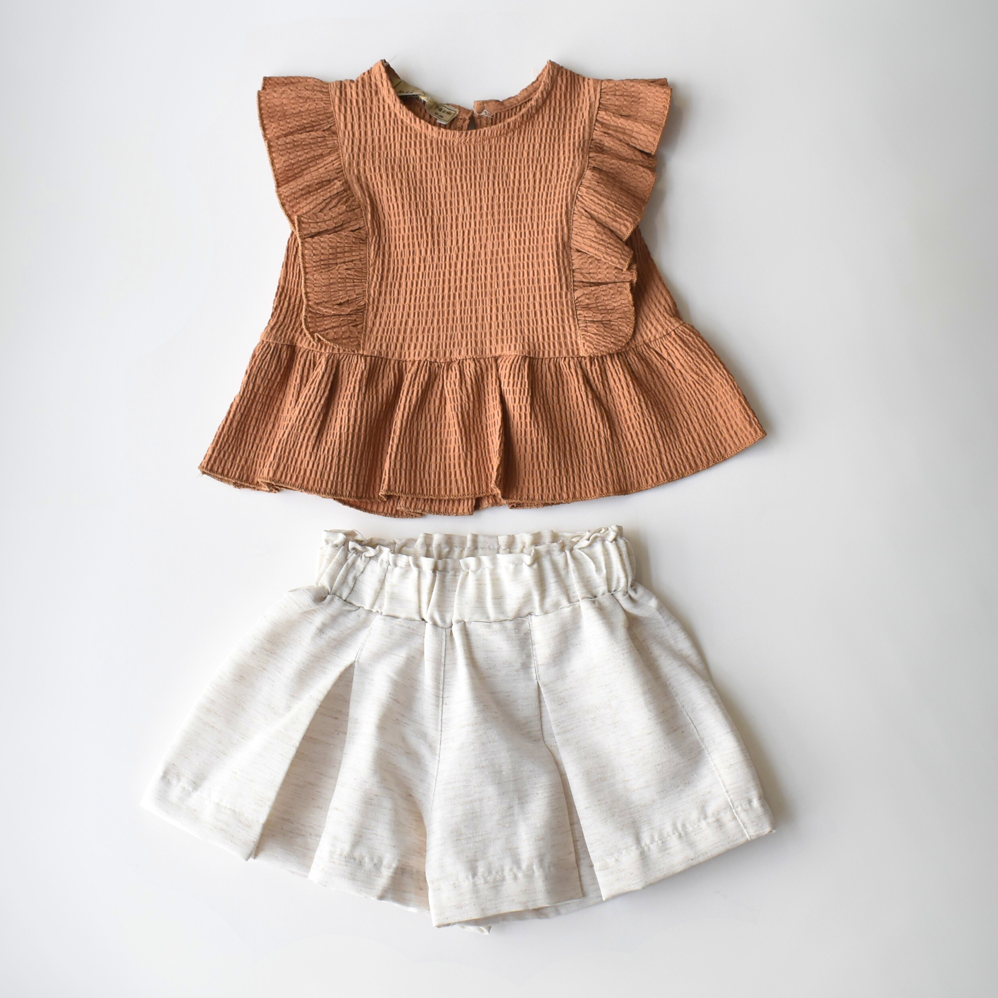 Sleeveless toddler outfit with a brown ruffle peplum top and high-waisted white pleated shorts, designed for casual summer wear.