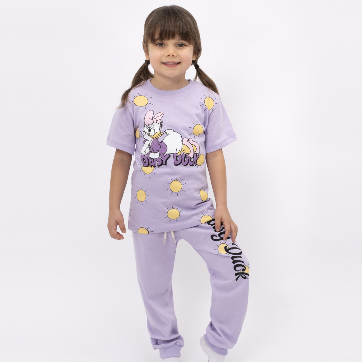 Smiling girl wearing a lilac Daisy Duck lounge set with short-sleeve graphic top and matching jogger pants decorated with yellow sun prints.