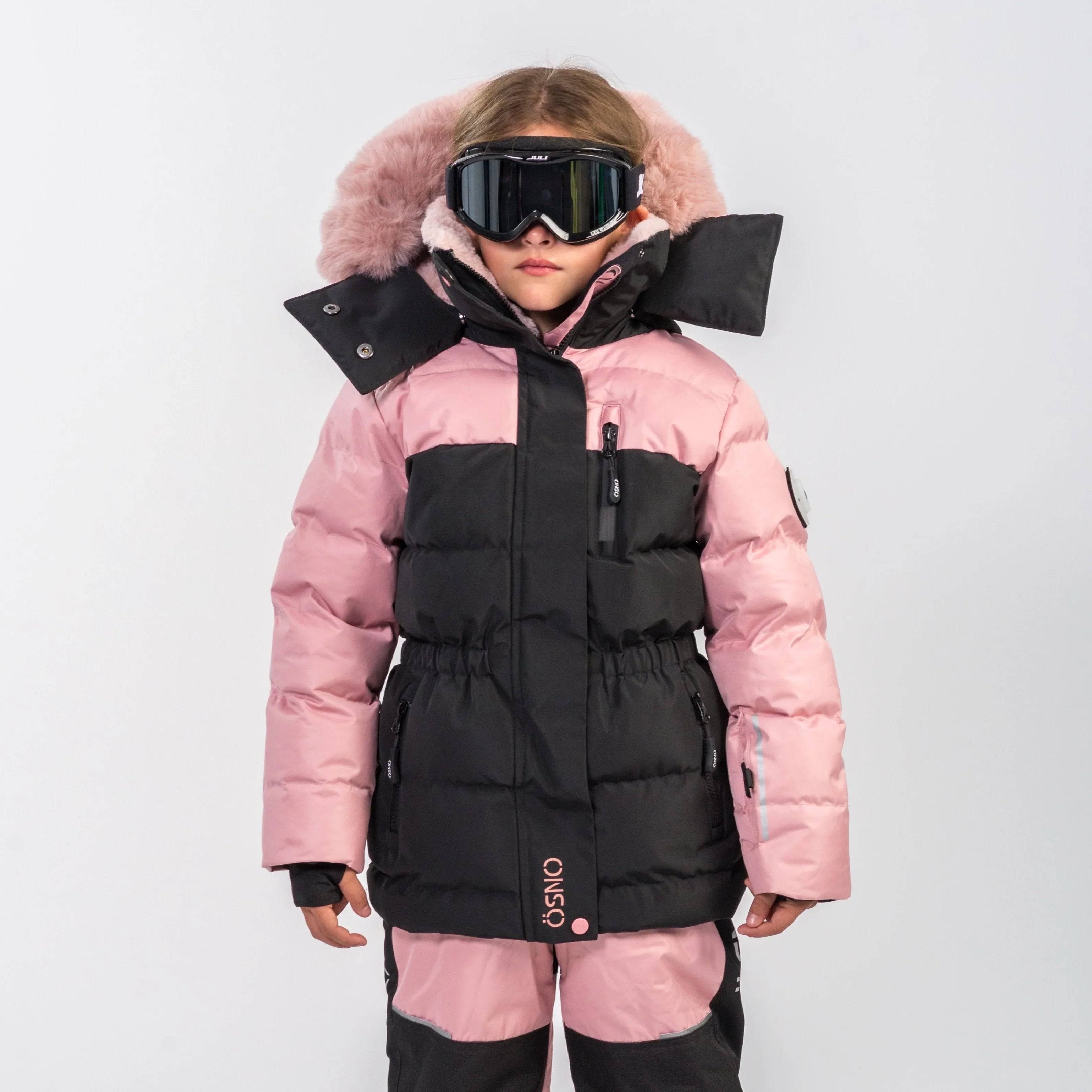 Girl in pink and black OSNO snowsuit jacket with fur-trimmed hood and ski goggles, pink snow pants