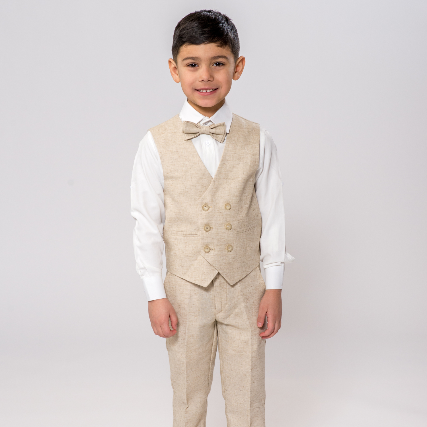 Young boy wearing a beige vest suit with matching trousers, white shirt and bow tie, smiling while standing against a plain background in formal kids attire