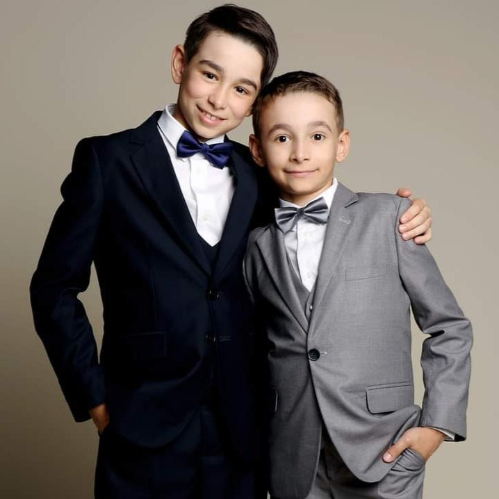 Two boys standing together in navy and grey formal suits with bow ties, styled for boys suits for weddings in Canada, showing coordinated looks for page boys or siblings.