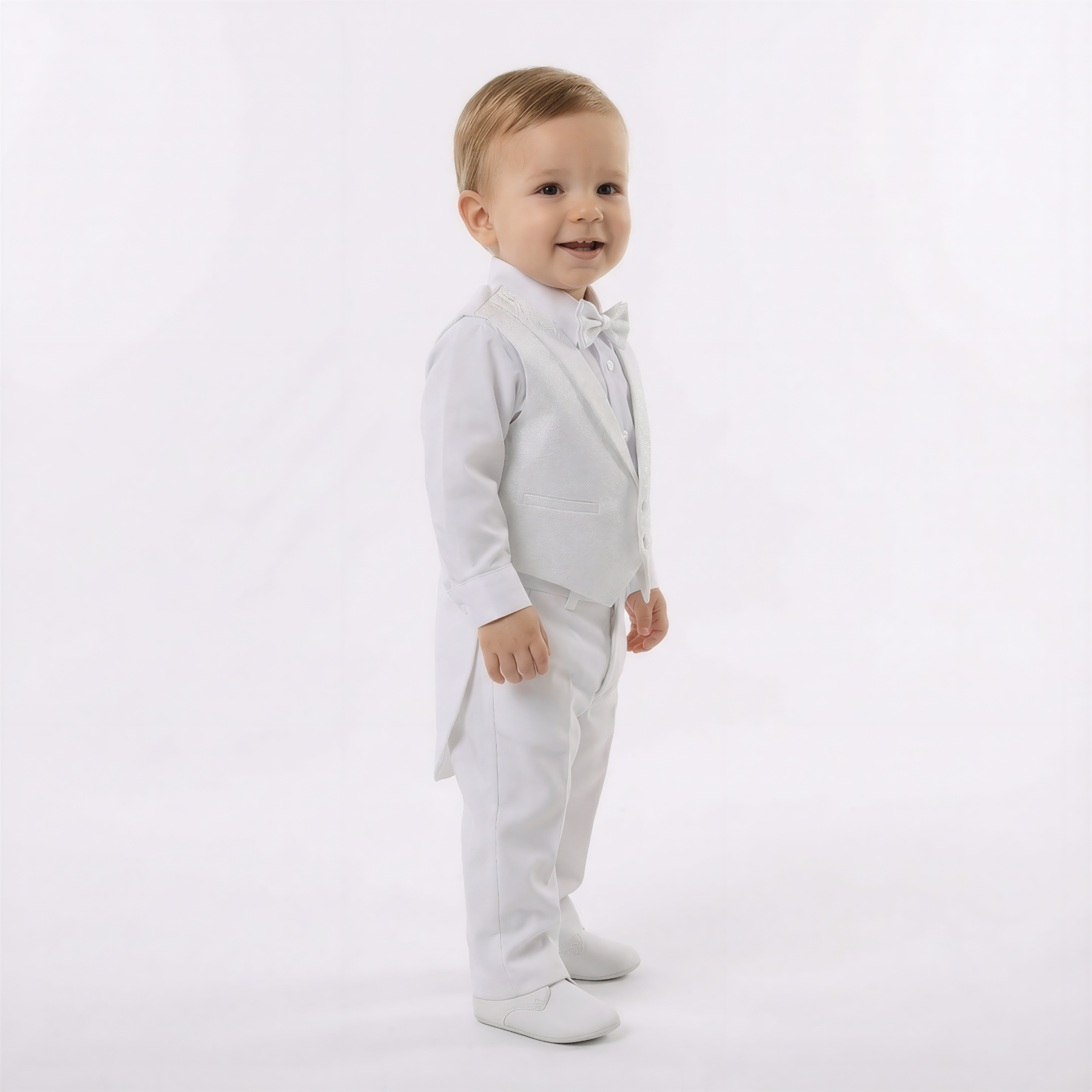 Toddler boy wearing a white formal suit with bow tie and vest, standing and smiling, dressed for a wedding or christening ceremony.