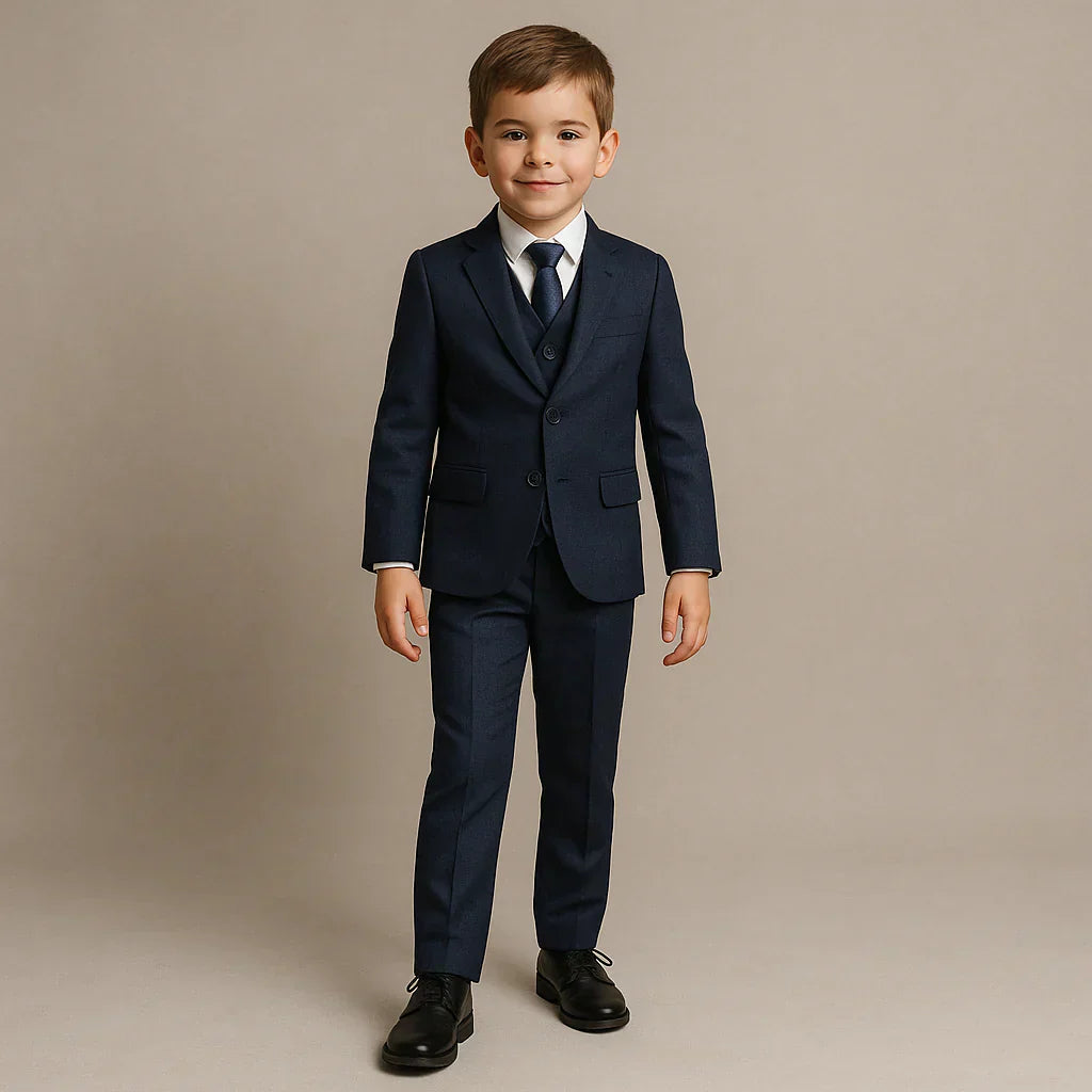 Young boy dressed in a navy blue suit with a matching tie and white shirt, standing and smiling confidently in front of a neutral beige background. 
