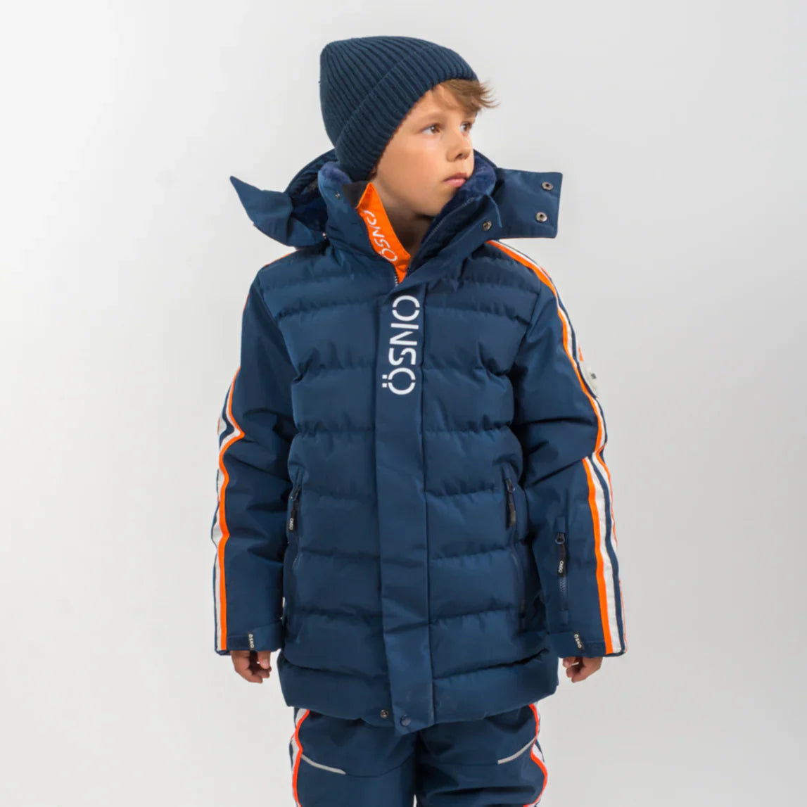 Boy wearing navy ÖSNO ski jacket with orange stripe detail and knit beanie.