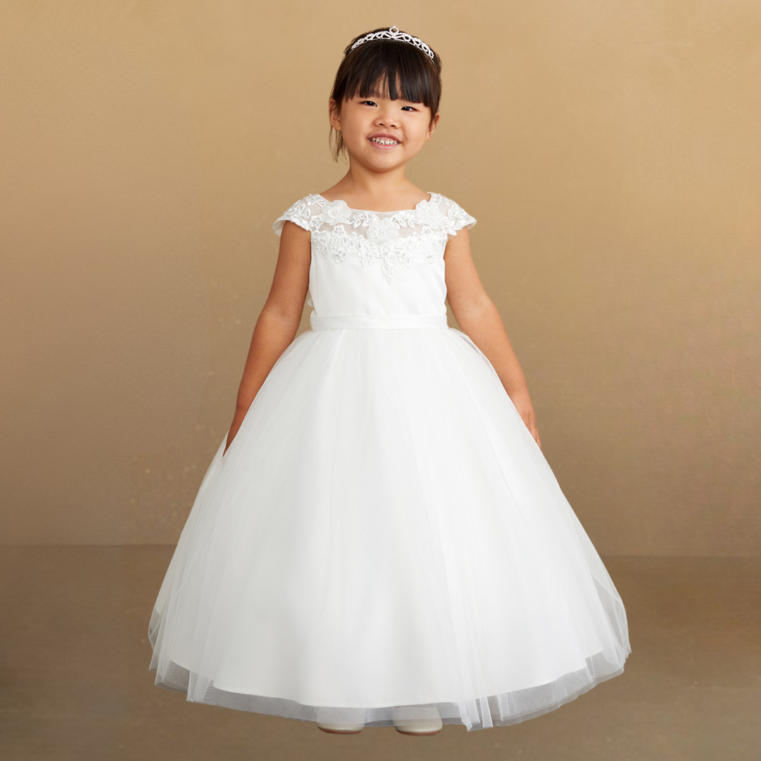 Smiling young girl in a floor-length white gown with a full tulle skirt, lace-embroidered bodice, cap sleeves, satin waistband & a sparkling tiara.