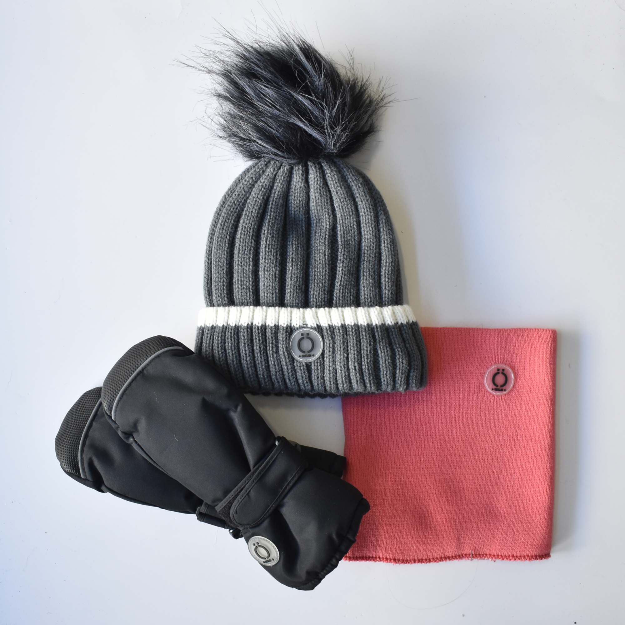 Three-piece kids’ winter set including a grey ribbed knit beanie with faux fur pom-pom, coral pink neck warmer & black insulated mittens with snaps.