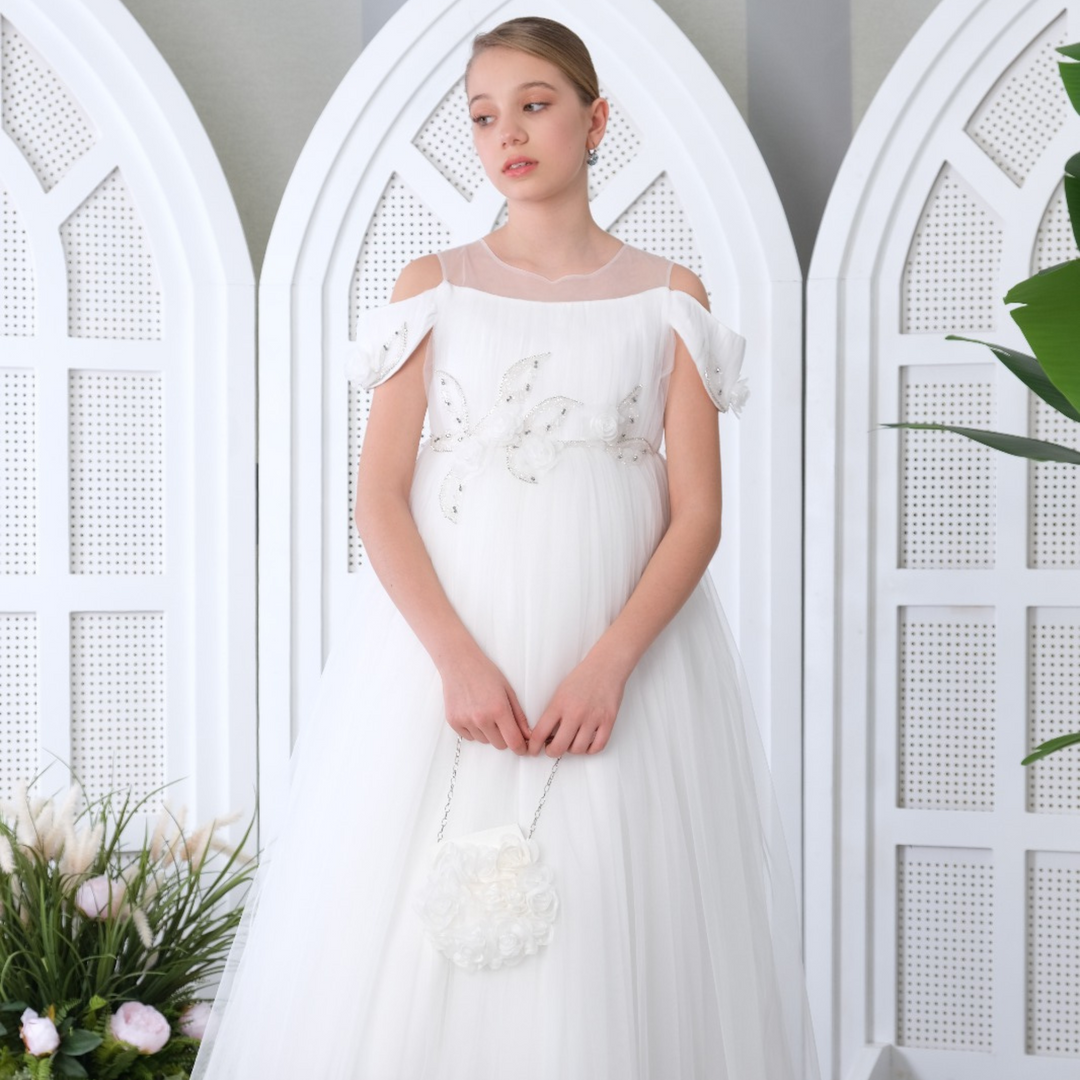 Girl in an elegant white communion gown with cold-shoulder sleeves, beaded floral waist detail, and tulle skirt, holding a white rosette purse.