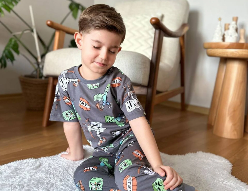 A boy kneeling on a white rug in a grey two-piece pyjama set with a green monster print.