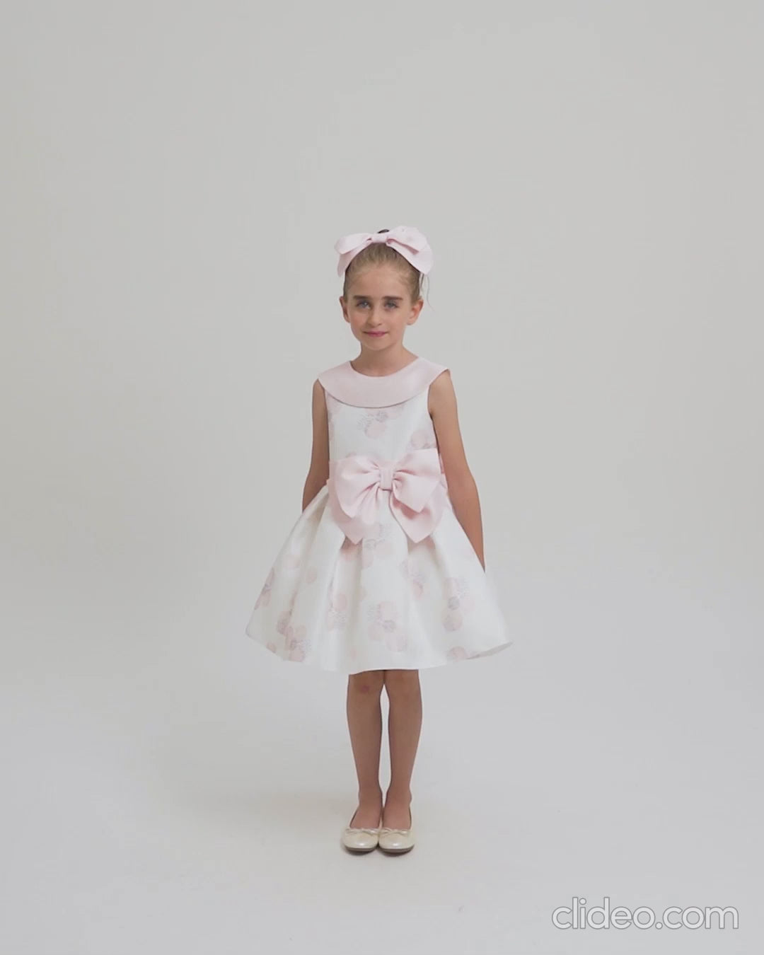 Girl wearing a white sleeveless dress with soft pink floral patterns and a large front bow, styled with matching headband in a white studio.