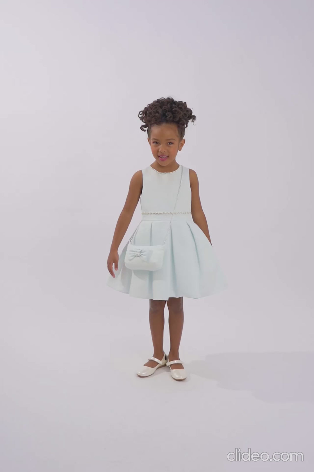 Girl wearing a sleeveless pastel blue dress with pearl trim and matching purse, standing in a white studio with neutral lighting and soft shadow.