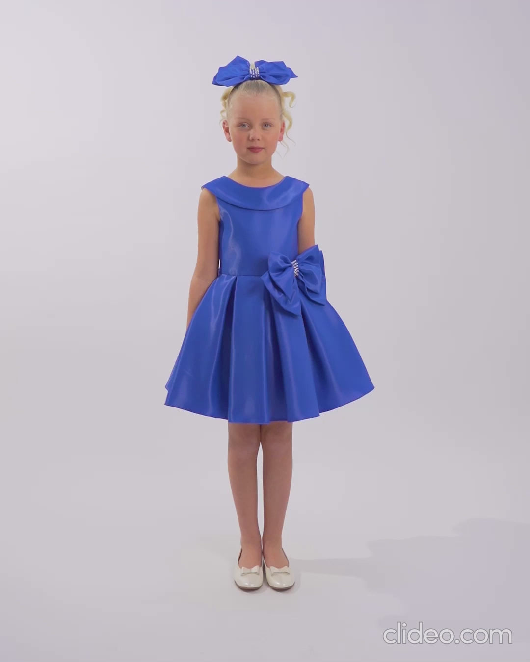 Girl in a bright royal blue sleeveless dress with large side bow and matching headband, standing poised against a plain white studio background.